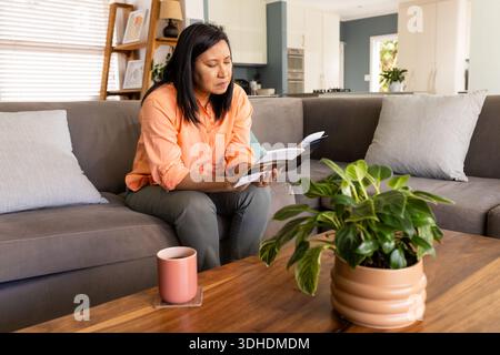 Femme asiatique d'âge moyen portant chemisier pêche assis sur un canapé gris lisant livre de poche à la maison Banque D'Images
