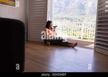 Femme mature assise sur le sol près de la porte du balcon, parlant dans le smartphone, tasse en céramique à côté d'elle Banque D'Images