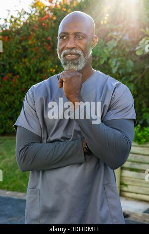Homme afro-américain senior debout dans le jardin portant des gommages gris et une montre-bracelet, lumière du soleil Banque D'Images