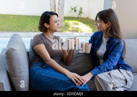 Diverses amies féminines assis près les uns des autres sur le canapé-terrasse tenant des tasses beiges assorties près de la piscine Banque D'Images