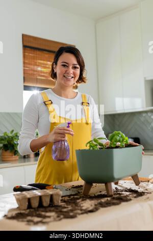 Femme afro-américaine adulte tendant dans la cuisine dans une combinaison jaune tenant la bouteille violette par le planteur Banque D'Images