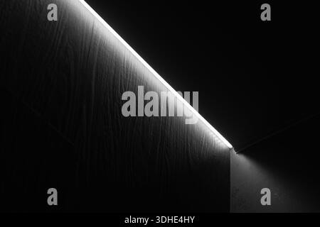 Une scène sombre et minimaliste présente une fine ligne lumineuse de plafond le long d'un mur blanc et un faisceau vers le bas qui crée un point doux et atmosphérique en dessous Banque D'Images
