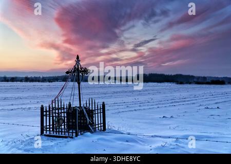 Paysage rural d'hiver au coucher du soleil. Un sanctuaire au bord de la route au premier plan. Spectaculaires nuages violets dans le ciel. Un champ couvert de neige. Podlachia, Polan Banque D'Images