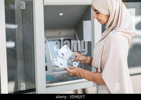 Heureux assez millénaire femelle islamique en hijab comptant beaucoup d'euros près de atm extérieur Banque D'Images