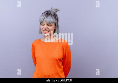 Jeune femme aux cheveux gris en pull orange souriant avec les yeux fermés sur un fond violet. Banque D'Images