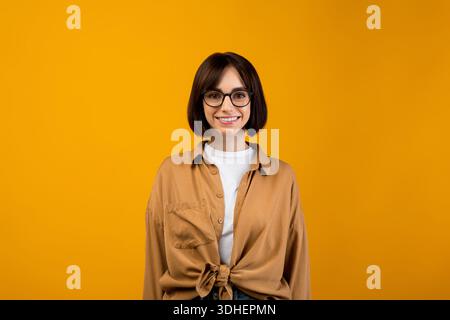 Étudiante joyeuse portant des lunettes et souriant à la caméra, posant sur fond jaune de studio Banque D'Images