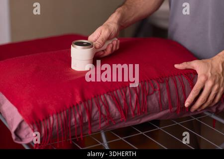 Foulard en laine rouge nettoyant les mains avec rouleau à peluches sur le tissu frangé, texture gros plan, éclairage chaud de la chambre à coucher, mouvement prudent, épilation d'animaux de compagnie Banque D'Images
