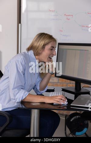 Femme mi-adulte dans la chemise rayée penchant, riant, en utilisant la souris au bureau avec le moniteur de feuille de calcul Banque D'Images