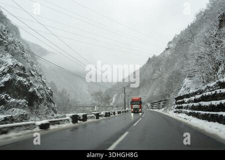 Un camion se déplace le long d'une route sinueuse entourée de montagnes enneigées. La faible visibilité et les nuages couvrent la scène alors que le temps hivernal affecte trav Banque D'Images