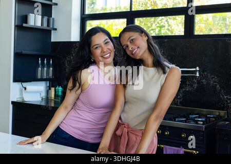 Deux amies se penchant sur l'îlot de la cuisine, souriant vers la caméra tout en portant des hauts sans manches Banque D'Images