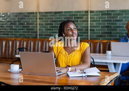 Femme africaine adulte travaillant sur un ordinateur portable argenté au café portant des écouteurs de chemisier jaune ordinateur portable au repos Banque D'Images