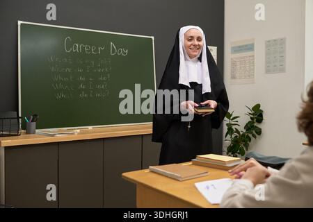 Femme caucasienne d'âge moyen habillée en nonne debout dans la salle de classe tenant des livres, souriant et s'engageant avec l'élève pendant le jour de la carrière, tableau noir avec des questions en arrière-plan Banque D'Images