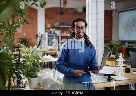 Portrait de jeune homme noir adulte souriant tout en tenant l'ordinateur portable dans le bureau Banque D'Images
