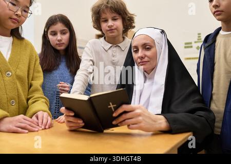 Nonne caucasienne d'âge moyen assise à table lisant la Bible tandis que groupe multiethnique d'enfants et d'adolescents debout autour de l'écoute attentive dans la salle de classe Banque D'Images