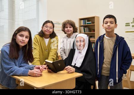 Religieuse caucasienne assise au bureau tenant la Bible entourée de divers groupes d'enfants et d'adolescents debout dans la salle de classe, tous regardant la caméra avec des expressions neutres Banque D'Images