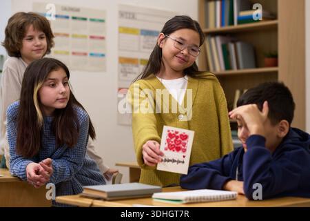 Groupe d'enfants multiethniques interagissant en classe, fille asiatique présentant une carte faite à la main avec des cœurs rouges au garçon assis à un bureau, deux autres filles observant et souriant à proximité Banque D'Images