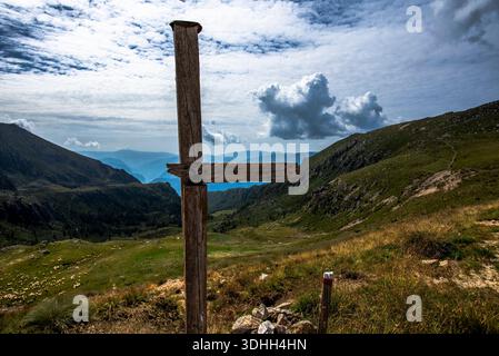 Croix en bois altérée debout sur un versant alpin surplombant des vallées verdoyantes et des sommets lointains de la chaîne de Lagorai dans le Trentin, en Italie. Banque D'Images