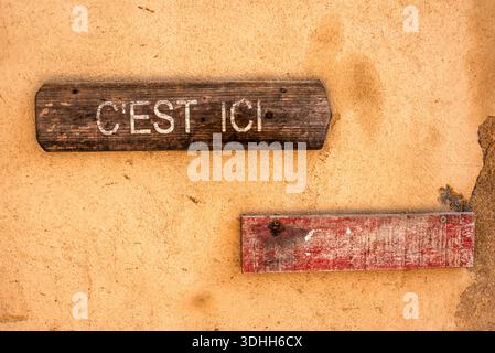 Panneau en bois vieilli lisant “C’est ici” sur un mur de plâtre rustique en Corse, France. Concept de direction, typographie minimale, texture méditerranéenne et Banque D'Images