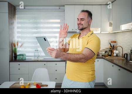 Homme moyen adulte appel vidéo à l'aide d'une tablette numérique dans la cuisine Banque D'Images