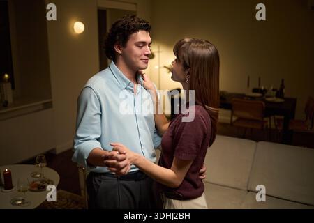 Jeune homme adulte caucasien et jeune femme adulte caucasienne dansant ensemble dans le salon, se tenant la main et se regardant, partageant un moment intime à l'intérieur Banque D'Images