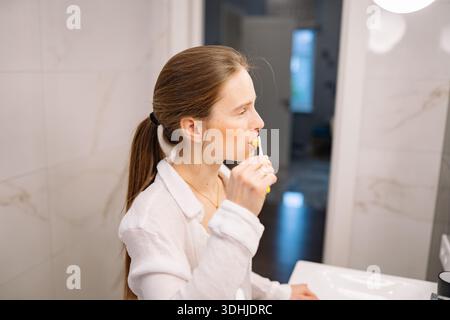 Jeune femme se tient dans la salle de bain, se brossant les dents tout en regardant son reflet. La lumière du matin remplit la pièce, créant une atmosphère lumineuse. Banque D'Images