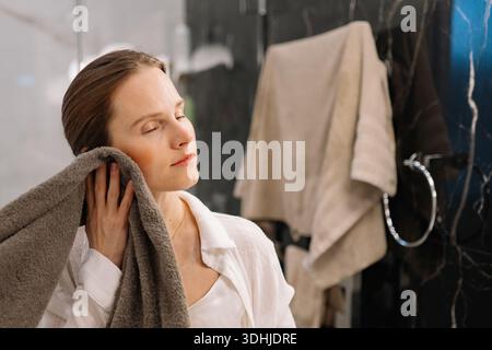 Une femme se tient dans une salle de bain après avoir pris une douche. Elle sèche ses cheveux avec une serviette tandis que les serviettes sont accrochées à des crochets en arrière-plan. La chambre a dar Banque D'Images