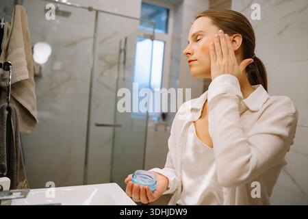 Une femme se tient devant un lavabo de salle de bain en appliquant un produit de soin de la peau sur son visage. La salle de bain au design moderne est baignée de lumière naturelle Banque D'Images