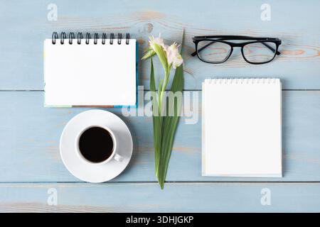 Un bureau en bois bleu avec des cahiers vierges, des verres, un stylo, du glaïeule et du café. Tasse de café du matin. Banque D'Images