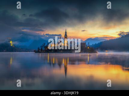 Vue aérienne de l'île avec l'église de l'Assomption reflétant dans les eaux tranquilles, sous un ciel vibrant au crépuscule, Spodnja Lua, Slovénie. Banque D'Images