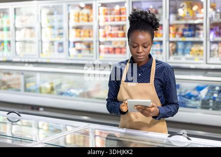 Une jeune femme employée d'une épicerie dans un tablier vérifie l'inventaire des aliments congelés et met à jour les niveaux de stock sur une tablette tout en se tenant dans l'allée réfrigérée Banque D'Images