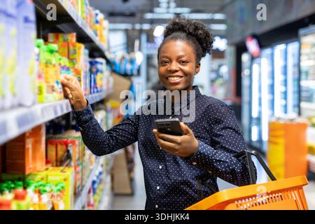 Une jeune femme dans un supermarché moderne utilise un smartphone pour vérifier une liste de courses tout en sélectionnant volontiers des produits d'épicerie frais et en plaçant des articles dans son panier Banque D'Images