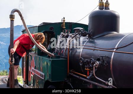 Brienzer Rothorn, Suisse - 26 août 2025 : une femme en chemise rouge travaillant sur une locomotive à vapeur vintage, ajustant l'équipement, versant de l'eau. Banque D'Images