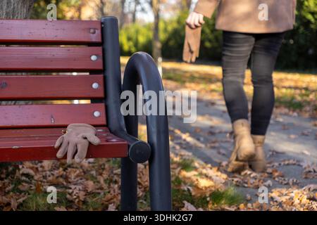 Vue rapprochée d'un seul gant beige laissé sur un banc rustique de parc à lattes, parsemé de feuilles d'automne brunes sèches, soulignant l'oubli, soli Banque D'Images