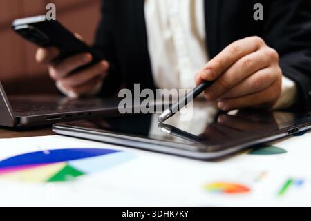 Homme d'affaires tient smartphone dans une main et stylet dans l'autre, interagissant avec la tablette tout en examinant les tableaux financiers et les graphiques sur les documents Banque D'Images