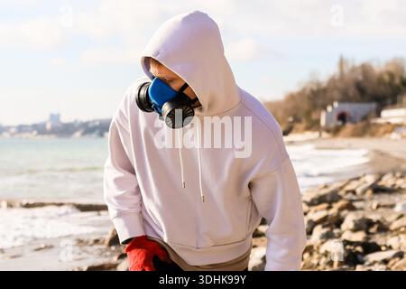 Individuel en tenue blanche, masque bleu, gants rouges regarde vers le bas sur la plage rocheuse avec l'océan en toile de fond Banque D'Images