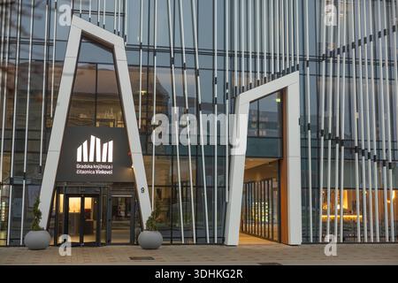 VINKOVCI, CROATIE, 12.13.2025:extérieur contemporain de la bibliothèque avec une entrée en voûte blanche frappante sertie dans des panneaux de verre. Éclairage intérieur chaleureux Banque D'Images