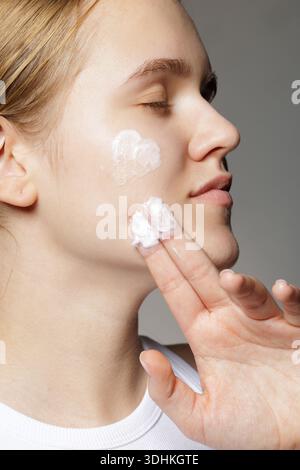 Profil en gros plan de la jeune femme appliquant la crème hydratante pour le visage sur fond gris. Idéal pour la bannière de site web de soins de la peau, blog de beauté, ou la clinique adverti Banque D'Images