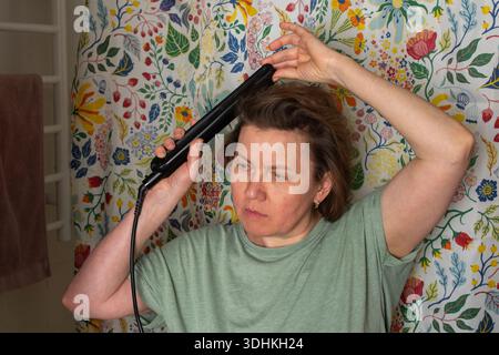 Une femme de 50 ans se tient dans une salle de bain avec un rideau de douche fleuri. Elle utilise un lisseur de cheveux, se concentrant sur ses cheveux tout en se préparant pour le Banque D'Images