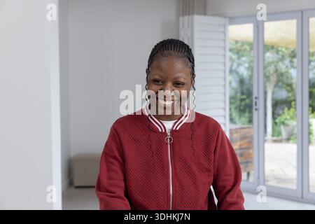 Femme afro-américaine debout et souriante à la maison par des portes coulissantes portant une veste rouge avec des tresses Banque D'Images