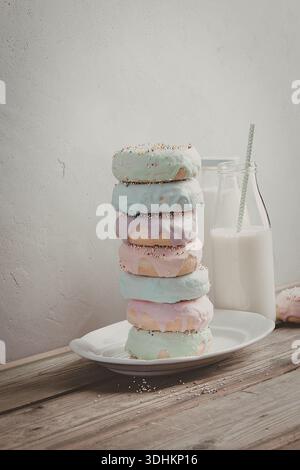 Une pile de six beignets de couleur pastel avec divers glaçons, accompagnés d'une bouteille de lait avec une paille, placée sur une table en bois rustique. Banque D'Images