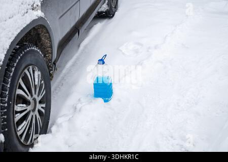 Réservoir de liquide lave-glace bleu debout dans la neige près de la roue de voiture en hiver, concept d'entretien de véhicule par temps froid Banque D'Images