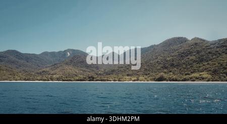 Une scène côtière tranquille avec un plan d'eau calme au premier plan et des montagnes verdoyantes en arrière-plan sous un ciel bleu clair. Banque D'Images