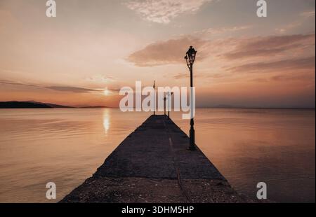 Une image sereine capturant un coucher de soleil sur des eaux calmes, avec une jetée en bois s'étendant dans l'eau. Le ciel est peint avec des teintes orange et rose, refl Banque D'Images