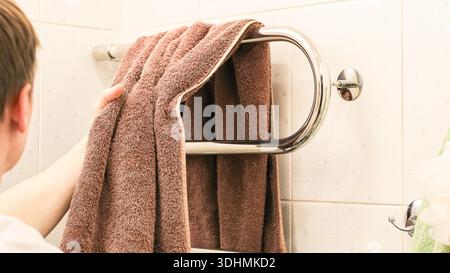 Vue arrière latérale d'un jeune homme prenant une serviette brune sur le rail chromé chauffant dans la salle de bains carrelée. Banque D'Images