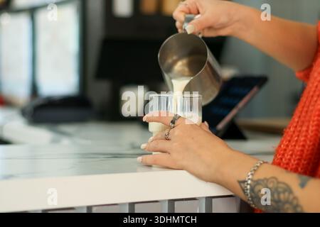 Gros plan des mains dans un chandail orange versant de la mousse de lait du pichet en métal dans un verre transparent sur le comptoir Banque D'Images