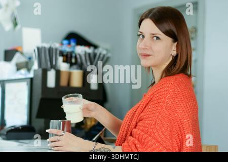 Jeune femme en pull orange sourit tout en tenant un petit verre de lait et une tasse au poste de travail du café Banque D'Images