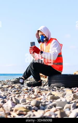 Homme en gilet orange et masque bleu tient le téléphone portable tout en s'agenouillant sur la côte rocheuse avec des vagues en vue. Banque D'Images