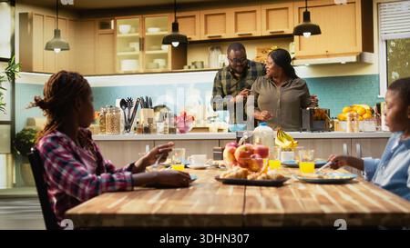 Parents et enfants à la maison servant le petit déjeuner dans la cuisine, mère préparant le repas au comptoir de la cuisine. Le moment familial capte la chaleur, la joie matinale et le fait d'être ensemble. Caméra B. Banque D'Images