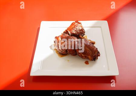 Ailes de poulet grillées BBQ avec glaçage sur une assiette blanche carrée sur fond rouge vif avec des ombres dures. Banque D'Images