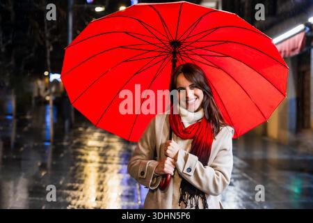 Femme souriante tenant un parapluie rouge pendant la pluie nocturne Banque D'Images